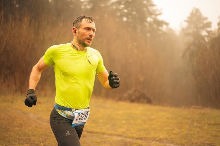 Wysportowany dorosły mężczyzna biegnący w biegu przełajowym w neonowej odzieży sportowej i rękawiczkach, uchwycony w ruchu na świeżym powietrzu.