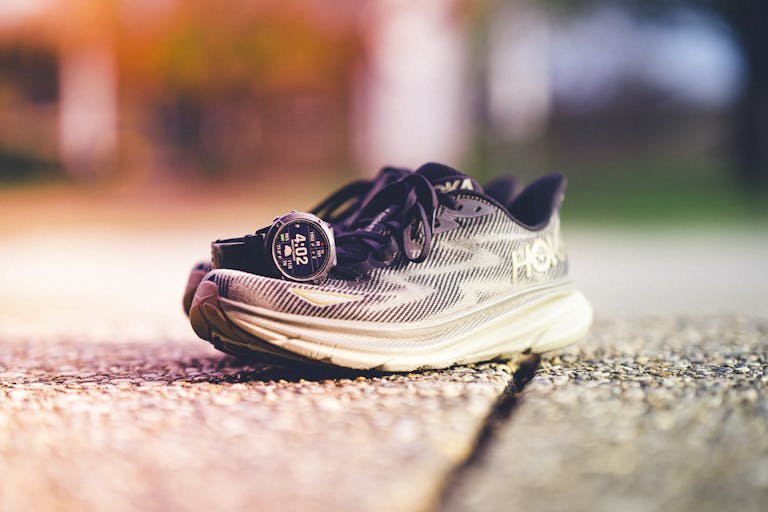 Para butów do biegania marki Hoka z zegarkiem Garmin obok nich na słonecznej bieżni na świeżym powietrzu.